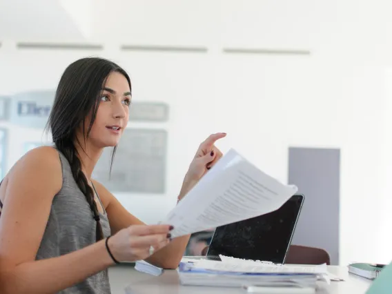 A woman holds a stack of papers next to a laptop computer.